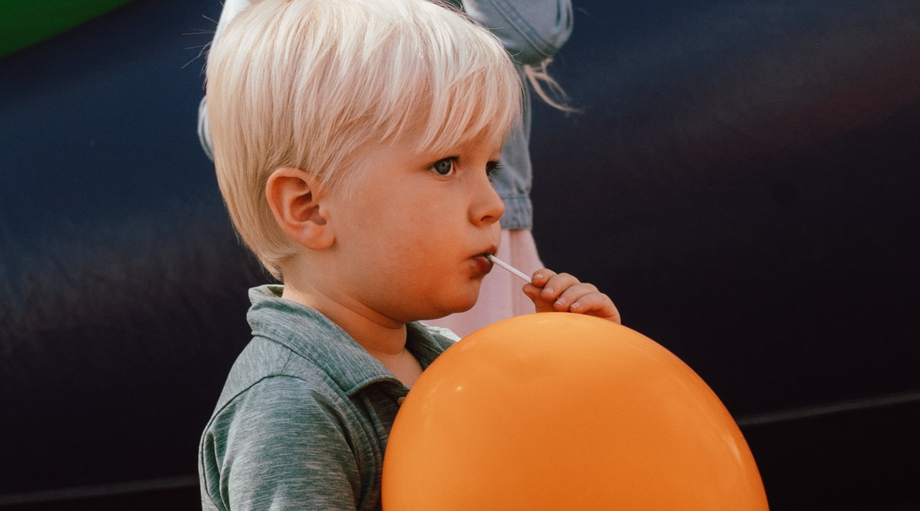 A little boy with a lollipop holding an orange balloon in front of the inflatables