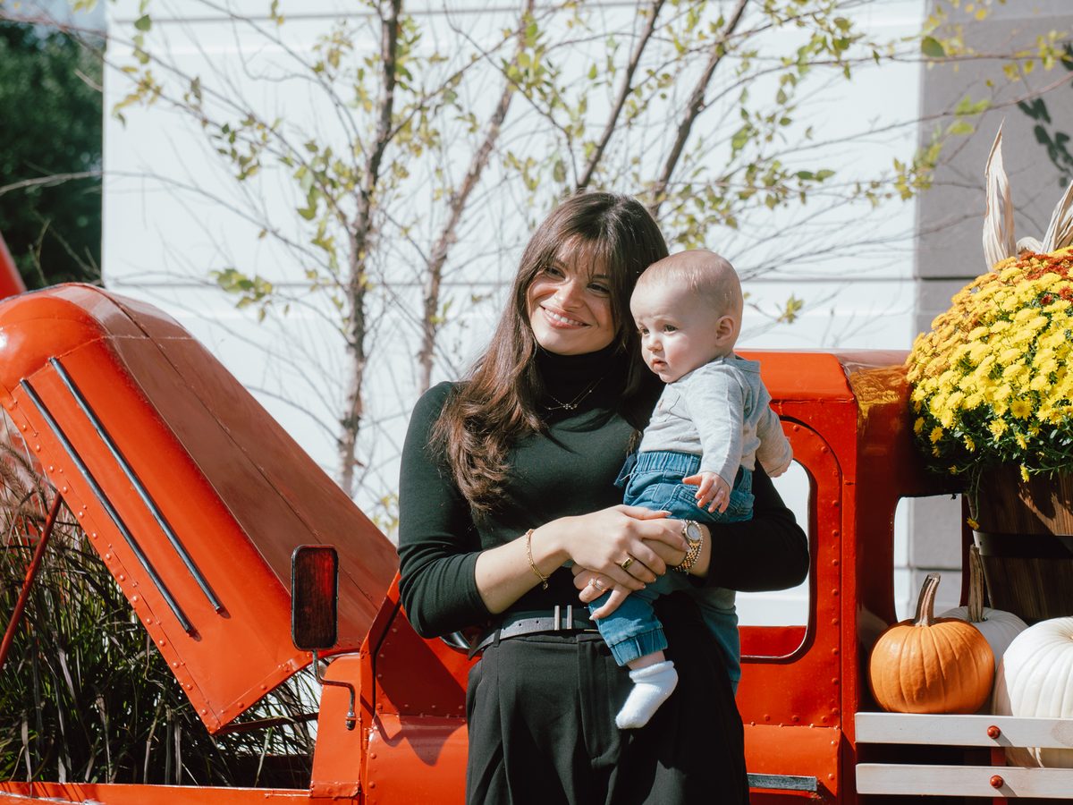 A mom holding her baby in front of the red truck, mums and pumpkins behind them